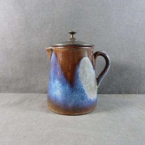 Puede incluir: Una jarra de cerámica con tapa. La jarra tiene un exterior marrón con detalles en azul y blanco. Tiene un asa y un pico. La tapa es de color metal oscuro. La jarra se utiliza probablemente para servir bebidas.