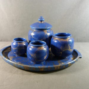 Könnte beinhalten: Ein blaues Keramik-Teeservice mit goldenen Akzenten. Das Set besteht aus einem Deckelgefäß, drei kleinen Töpfen und einem Tablett. Die blaue Glasur ist mit goldenen Wirbeln verziert. Das Tablett hat Griffe.