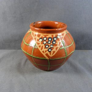 May include: A small, round, terracotta-colored ceramic pot with a wide opening. The pot is decorated with a geometric pattern of green lines and a central design featuring beige shapes and clusters of blue and white dots. The pot has a rustic, handmade appearance.