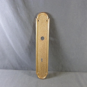 Op de afbeelding: Een gouden metalen deurplaat met een sleutelgat en een rond gat voor een deurklink. De plaat heeft een getextureerd oppervlak en een decoratief ontwerp.