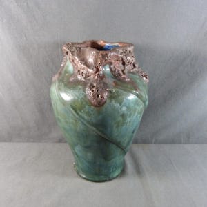 May include: A handcrafted ceramic vase with a unique design. The vase features a teal green glaze with brown textured accents around the rim. The vase has a bulbous shape with a swirling pattern. A decorative piece for the home.