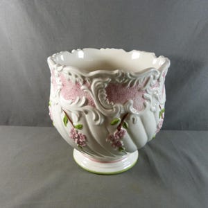 Peut inclure: Pot de fleurs en céramique blanche avec un bord festonné et un socle. Le pot présente des motifs floraux et de volutes roses, avec des accents verts sur les feuilles et les tiges. La base a une bordure verte.