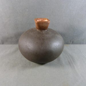 Könnte beinhalten: Eine runde, dunkelbraune Keramikvase mit einem quadratischen, kupferfarbenen Aufsatz. Die Vase hat eine glatte, matte Oberfläche und eine kugelförmige Form. Der Aufsatz hat eine kontrastierende Farbe und Textur.