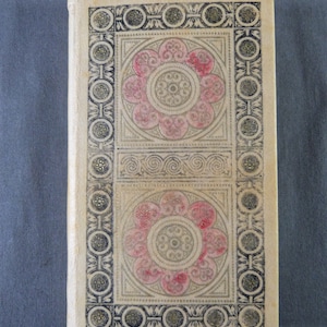 Puede incluir: Una cubierta de libro vintage con fondo blanco y un borde decorativo. El borde es negro y dorado con un patrón repetitivo de círculos y espirales. El centro de la cubierta presenta dos diseños florales rosas y blancos.