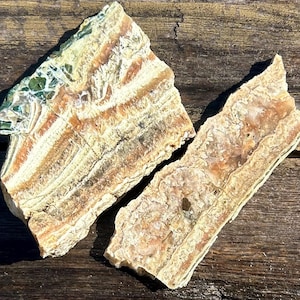 May include: Two raw, natural stone slabs with a layered, banded appearance. The stones feature a mix of colors, including beige, cream, orange, and green. The rough texture and irregular shapes suggest they are unpolished and in their natural state.