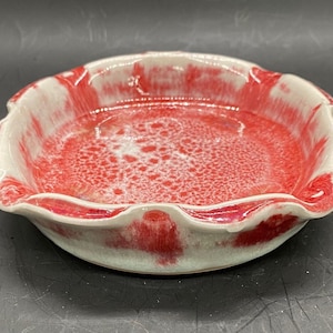 May include: A small, white ceramic dish with a scalloped edge. The dish is decorated with a red and white glaze that creates a marbled effect.