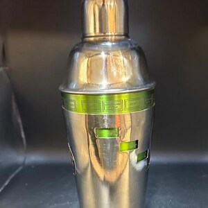 May include: A stainless steel cocktail shaker with a rounded top and a cylindrical body. The shaker features a green band with text and rectangular cutouts. The metal surface reflects the surrounding environment.