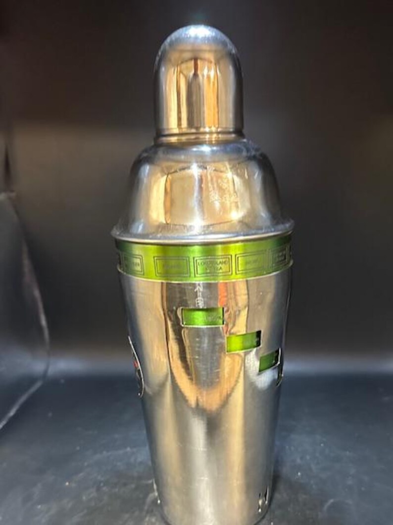 May include: A stainless steel cocktail shaker with a rounded top and a cylindrical body. The shaker features a green band with text and rectangular cutouts. The metal surface reflects the surrounding environment.