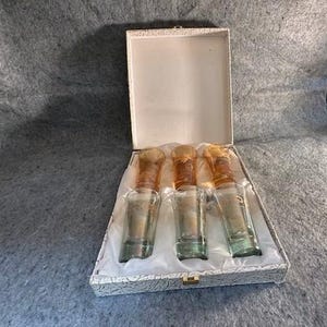 May include: A set of three clear glass perfume bottles with amber-colored stoppers, presented in a decorative box. The bottles are displayed on a white satin lining within a textured, off-white box with a gold clasp. The stoppers have a textured design.