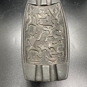 May include: Black ceramic ashtray with a carved design of stylized figures. The ashtray has a rectangular shape with rounded edges and three indentations on the top.
