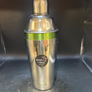 May include: A stainless steel cocktail shaker with a rounded top and a cylindrical body. A green band with cocktail names encircles the shaker. The text "Martinis + Merlot Handmade in India" is on a sticker on the front.