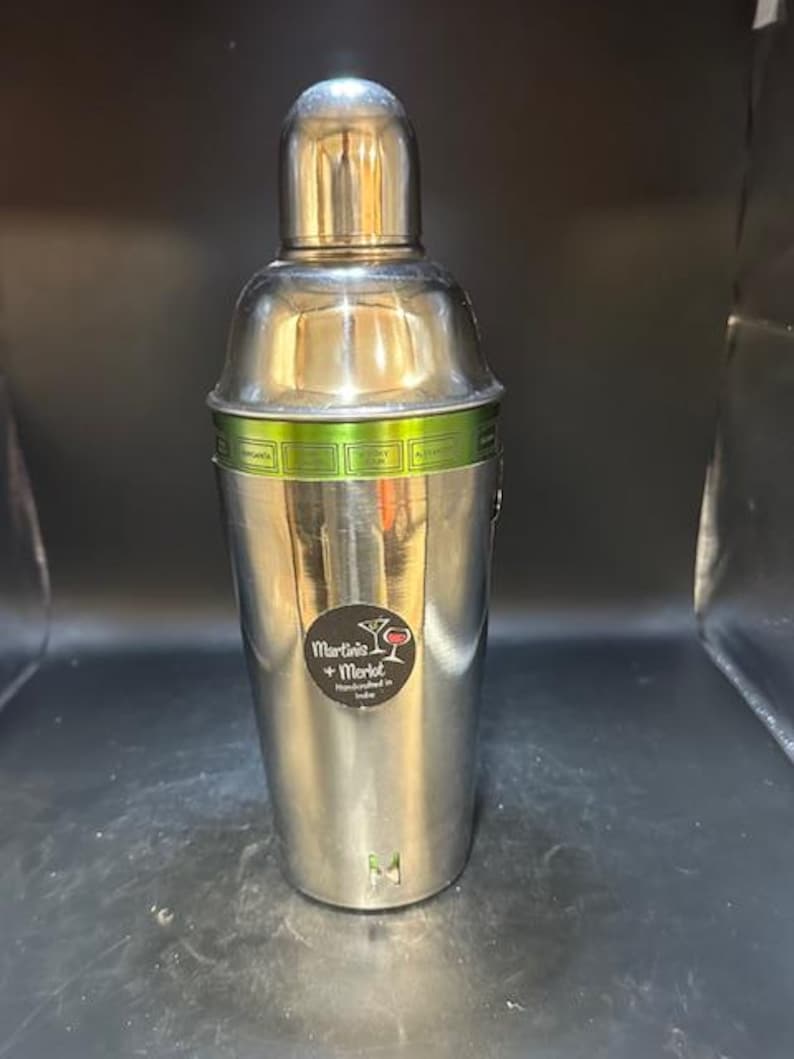 May include: A stainless steel cocktail shaker with a rounded top and a cylindrical body. A green band with cocktail names encircles the shaker. The text "Martinis + Merlot Handmade in India" is on a sticker on the front.