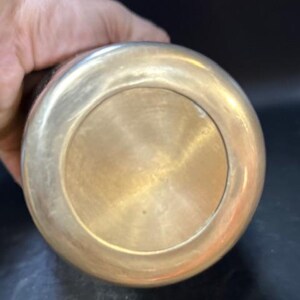 May include: A brass-colored metal object with a rounded base and a contrasting dark upper portion. The base has a circular design with a polished finish. The object is held by a hand, suggesting its size and portability. The item appears to be a decorative or functional piece.