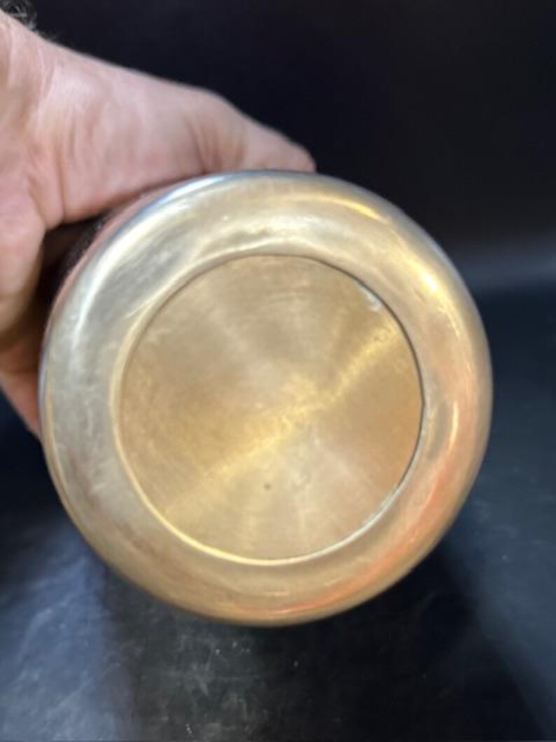 May include: A brass-colored metal object with a rounded base and a contrasting dark upper portion. The base has a circular design with a polished finish. The object is held by a hand, suggesting its size and portability. The item appears to be a decorative or functional piece.