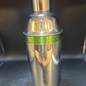May include: A stainless steel cocktail shaker with a shiny, metallic finish. The shaker has a rounded top and a cylindrical body. A green band with text is wrapped around the middle. The shaker is designed for mixing drinks.