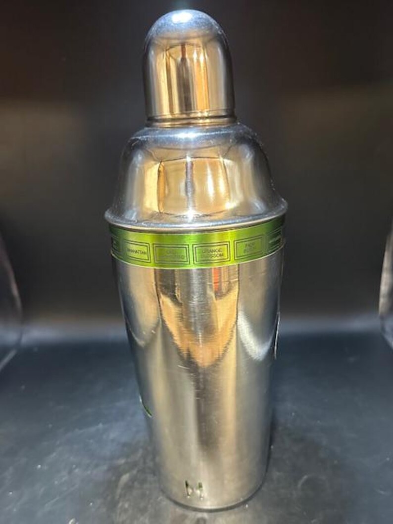May include: A stainless steel cocktail shaker with a shiny, metallic finish. The shaker has a rounded top and a cylindrical body. A green band with text is wrapped around the middle. The shaker is designed for mixing drinks.
