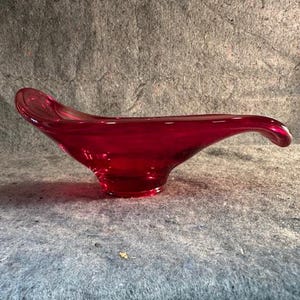 May include: A vibrant red glass bowl with a unique, flowing design. The bowl has a wide, shallow base that curves upwards to a pointed tip. The glass is transparent, allowing light to pass through, and is likely used for decorative purposes.
