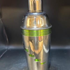 May include: A stainless steel cocktail shaker with a shiny, metallic finish. The shaker has a rounded top and a cylindrical body with rectangular cutouts revealing a green interior. A green band with text wraps around the middle.