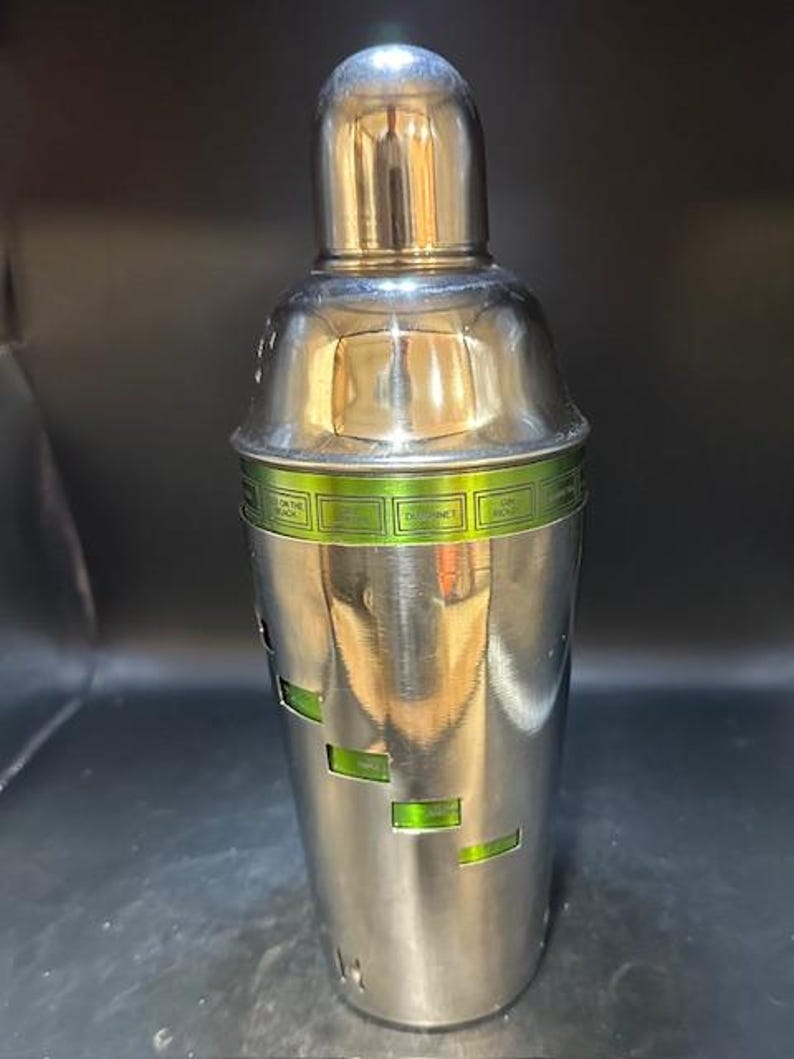 May include: A stainless steel cocktail shaker with a shiny, metallic finish. The shaker has a rounded top and a cylindrical body with rectangular cutouts revealing a green interior. A green band with text wraps around the middle.