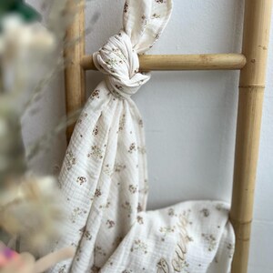 Peut inclure: Une couverture en mousseline de coton blanche avec un motif floral et le nom "Louella" brodé en fil brun.