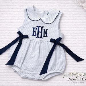 May include: A sleeveless white and light blue pinstriped romper featuring a rounded collar with dark blue trim, a dark blue embroidered monogram with the letters EHN, and two dark blue ribbon ties attached at the waist on each side.