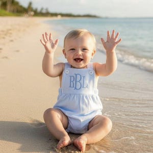 Könnte beinhalten: Ein ärmelloser Strampler in Hellblau mit geraffter Taille und zwei weißen Knöpfen an den Schulterträgern. Die Vorderseite des Kleidungsstücks ist mit den Monogrammbuchstaben RBL in passendem hellblauem Garn in klassischer Schreibschrift bestickt.