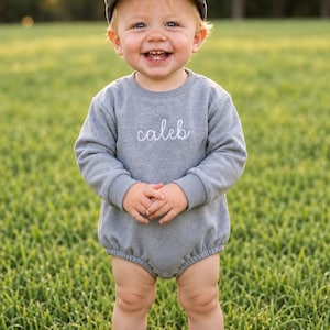 Könnte beinhalten: Ein grauer Langarm-Strampler mit Rundhalsausschnitt und gerippten Bündchen, der auf der Brust den in weißer Schreibschrift gestickten Namen "caleb" trägt. Das Kleidungsstück wird mit einer passenden grauen Baseballkappe kombiniert.