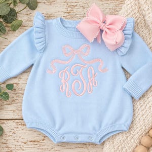 May include: A light blue long-sleeve knit romper featuring ruffled shoulders and a pink embroidered monogram with a bow design above the letters. A large pink hair bow with white scalloped edges is attached to the upper right shoulder area.