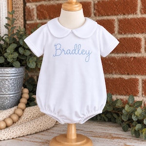 May include: A white short-sleeved bubble romper featuring a rounded Peter Pan collar and the name Bradley embroidered in light blue script across the chest. The garment is displayed on a wooden mannequin stand, showcasing a clean, simple design.