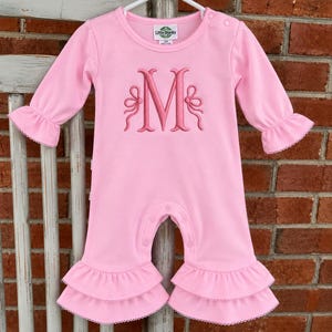 May include: A light pink long-sleeve romper featuring a dark pink embroidered letter M monogram flanked by two decorative bows. The garment has a round neckline, snap closures at the shoulder and crotch, and double-layered ruffled leg cuffs.