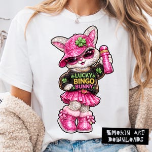 May include: White t-shirt featuring a cartoon bunny wearing a pink hat, sunglasses, and a tutu. The bunny holds a pink object and the shirt reads "LUCKY BINGO BUNNY". The design includes shamrocks and a heart.