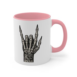 Puede incluir: Taza de cer&aacute;mica blanca con interior y asa rosas. La taza presenta una ilustraci&oacute;n en blanco y negro de una mano esquel&eacute;tica haciendo el gesto de rock and roll, con el nombre "OZZY" escrito en los nudillos.