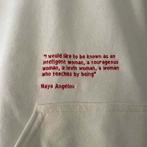 May include: White hoodie with a red embroidered quote by Maya Angelou: "I would like to be known as an intelligent woman, a courageous woman, a loving woman, a woman who teaches by being."
