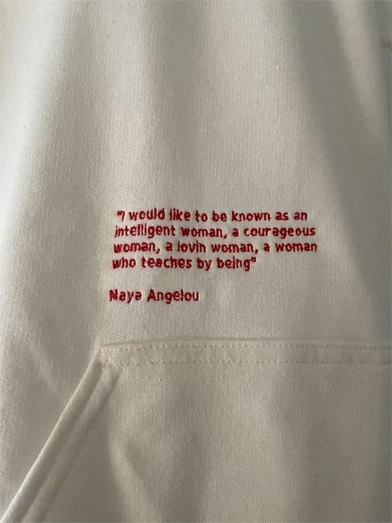 May include: White hoodie with a red embroidered quote by Maya Angelou: "I would like to be known as an intelligent woman, a courageous woman, a loving woman, a woman who teaches by being."