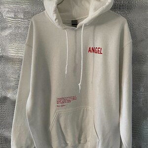 May include: White hooded sweatshirt with a red embroidered "ANGEL" on the front and a red embroidered quote on the pocket. The quote reads: "I would like to be known as an intelligent woman, a courageous woman, a woman who dares to be a woman who teaches by being."