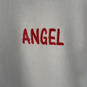 May include: White sweatshirt with the word "ANGEL" embroidered in red.