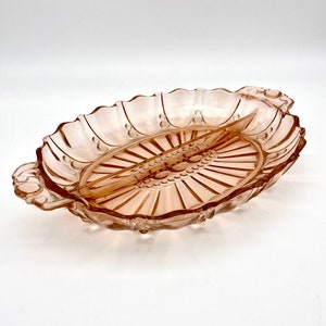 Puede incluir: Un plato de servir de vidrio rosa dividido con un borde festoneado y un patrón decorativo. El plato tiene dos asas y un divisor central elevado.