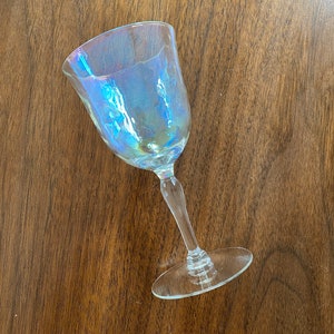 K&ouml;nnte beinhalten: Ein klares, schillerndes Weinglas mit rundem Kelch und langem Stiel. Das Glas hat einen subtilen Regenbogen-Schimmer, der verschiedene Farben reflektiert. Das Glas steht auf einer dunkelbraunen Holzoberfl&auml;che.