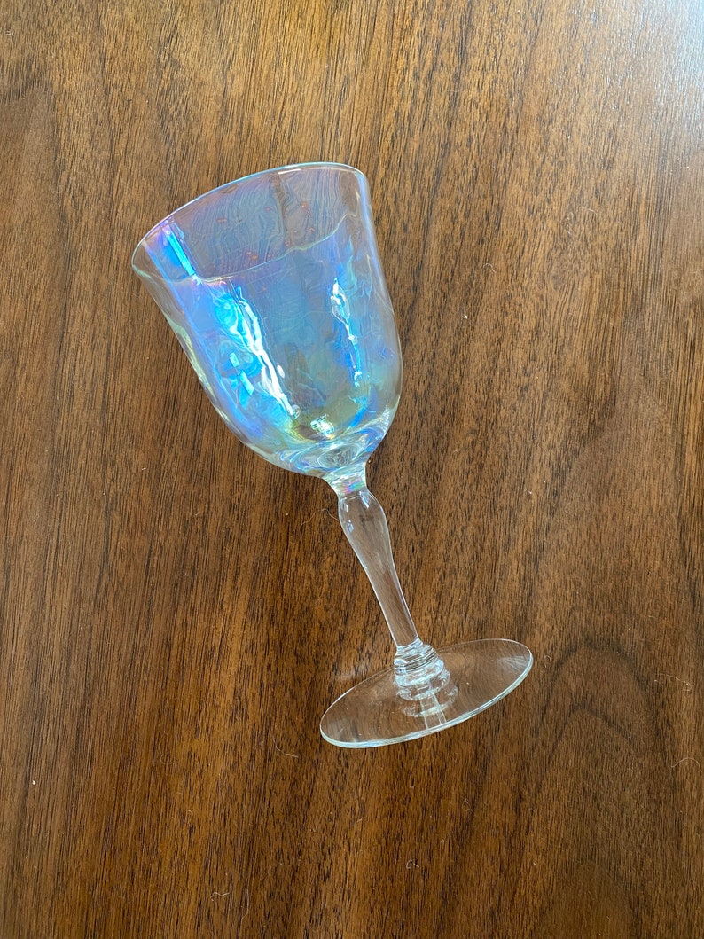K&ouml;nnte beinhalten: Ein klares, schillerndes Weinglas mit rundem Kelch und langem Stiel. Das Glas hat einen subtilen Regenbogen-Schimmer, der verschiedene Farben reflektiert. Das Glas steht auf einer dunkelbraunen Holzoberfl&auml;che.