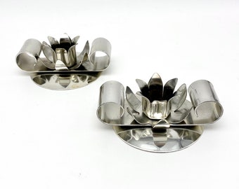 May include: Two silver candle holders with a flower design. Each holder has a round base and a decorative flower-shaped candle cup.