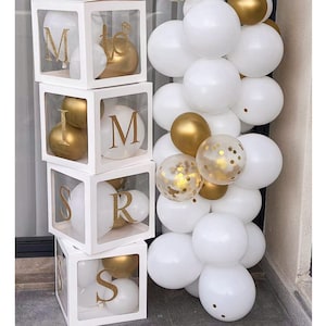May include: Four clear acrylic boxes stacked on top of each other, each box contains gold and white balloons. The boxes are decorated with the letters "MRS" in gold glitter. A garland of white and gold balloons is to the right of the boxes.