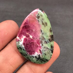 Op de afbeelding: Een gepolijste, druppelvormige zoisiet edelsteen met een opvallend centraal gedeelte van levendig magenta-roze robijn, omringd door gevlekt lichtgroen zoisiet en verspreide donkere zwarte minerale insluitingen over het gehele oppervlak.