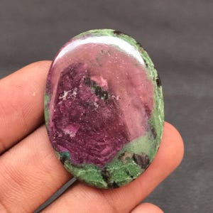 May include: An oval-shaped polished ruby zoisite gemstone featuring a prominent deep magenta center surrounded by mottled light green and dark black mineral inclusions, showcasing a smooth, reflective surface with natural color variations.