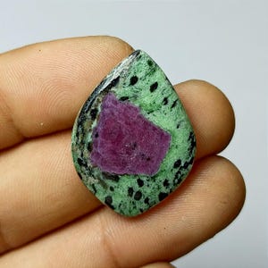 May include: A teardrop-shaped gemstone featuring a central, deep magenta-colored area surrounded by a speckled green field. The green area is dotted with black specks, and the edges are a dark, glossy color. The gemstone is held in a hand.