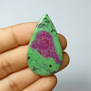 Może przedstawiać: Wypolerowany kamień szlachetny w kształcie łzy, prezentujący żywą kombinację kolorów. Kamień ukazuje centralny obszar w głębokiej magencie, otoczony nakrapianą, jasnozieloną matrycą. Małe czarne inkluzje są rozproszone.