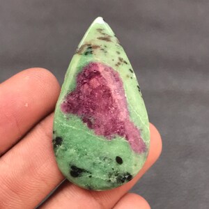 May include: A teardrop-shaped polished gemstone featuring a vibrant magenta center surrounded by a light green base with scattered black mineral inclusions. The stone has a smooth, rounded surface and a pointed top, displaying natural color variations.