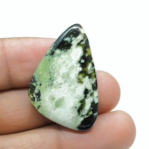 May include: A polished gemstone with a unique pattern. The stone features a mix of light green, white, and dark green colors. The shape is a rounded triangle, with a glossy, smooth surface. The gemstone is held in a hand.