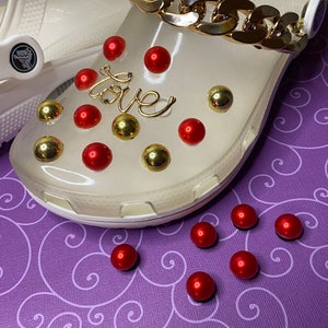 May include: White clogs with a gold chain and red and gold embellishments. The word "love" is spelled out in gold on the shoe.  There are several red and gold embellishments on the shoe and on a purple surface.
