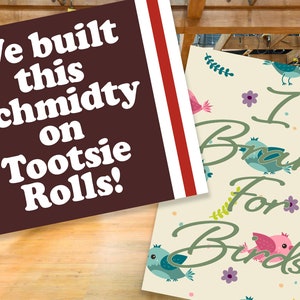 May include: Two signs with text on them. The first sign is brown with white text that says "We built this Schmidty on Tootsie Rolls!" The second sign is cream colored with green text that says "I Brake For Birds!" and has a pattern of colorful birds and flowers.