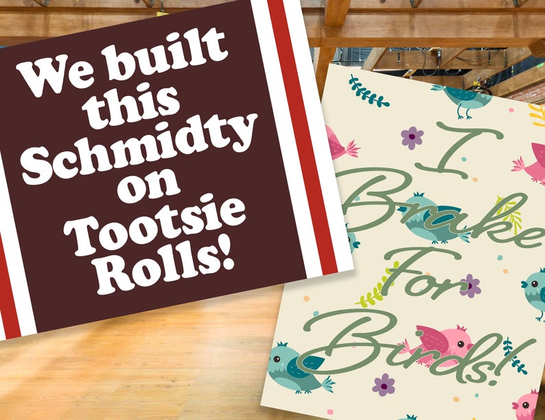 May include: Two signs with text on them. The first sign is brown with white text that says "We built this Schmidty on Tootsie Rolls!" The second sign is cream colored with green text that says "I Brake For Birds!" and has a pattern of colorful birds and flowers.